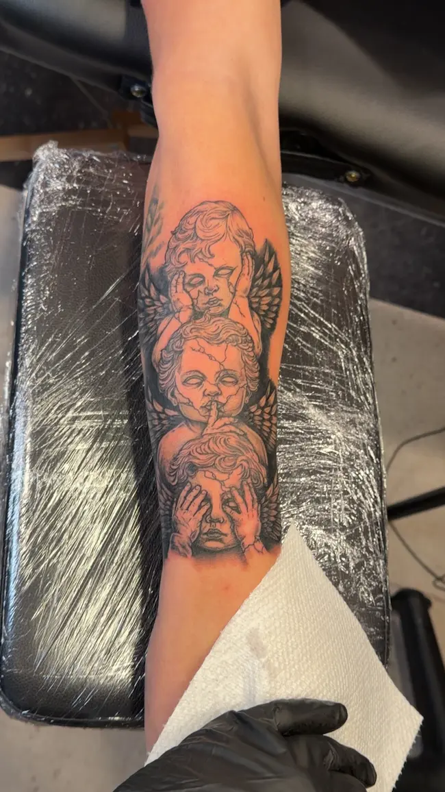 A vertical black and grey tattoo on a forearm featuring three winged cherubs with a cracked stone effect. They represent hear, speak, and see no evil by covering their ears, mouth, and eyes. The design has detailed shading and ancient statue vibes.